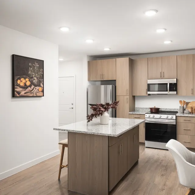 A modern kitchen featuring wooden cabinets, stainless steel appliances, and a central island. A painting of fruit hangs on the wall, and there are small kitchen appliances and utensils on the counters.