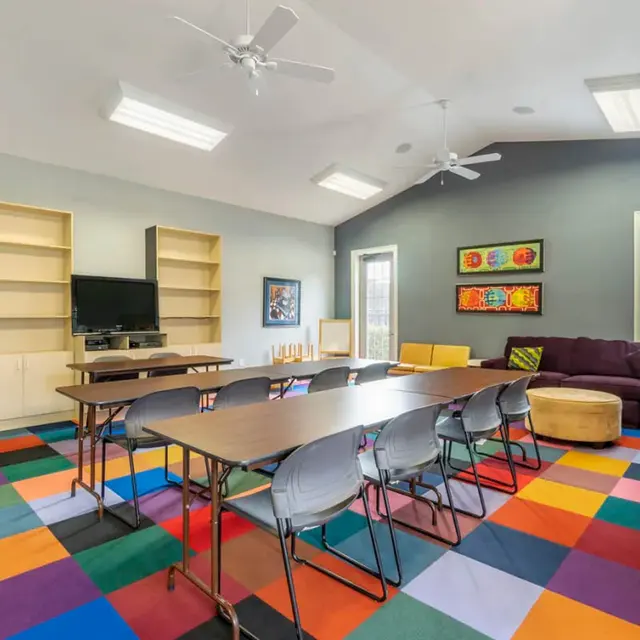 A brightly colored meeting room featuring a multi-colored floor, a long table with chairs, a couch, and shelves with a television.