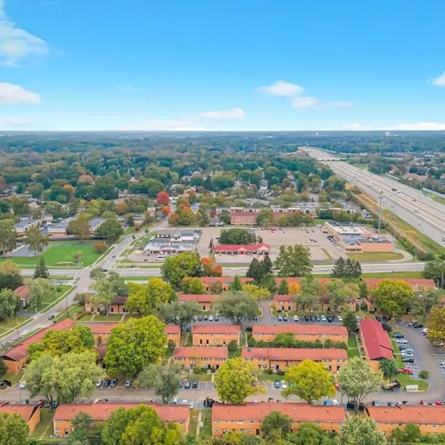 Aerial view of a suburban area featuring residential complexes, green trees, and a highway in the background.