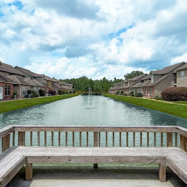 A serene view of a pond surrounded by residential buildings under a cloudy sky. A wooden bench is in the foreground, inviting relaxation by the water.
