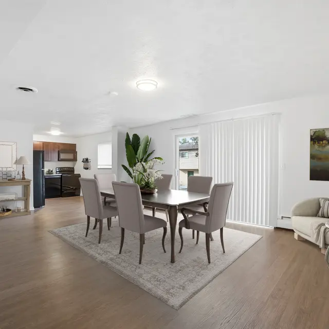 A spacious and modern dining area featuring a wooden dining table surrounded by six upholstered chairs, a large indoor plant, and a beige area rug. The room has large windows with white blinds allowing natural light in, along with light hardwood flooring and minimalist decor.