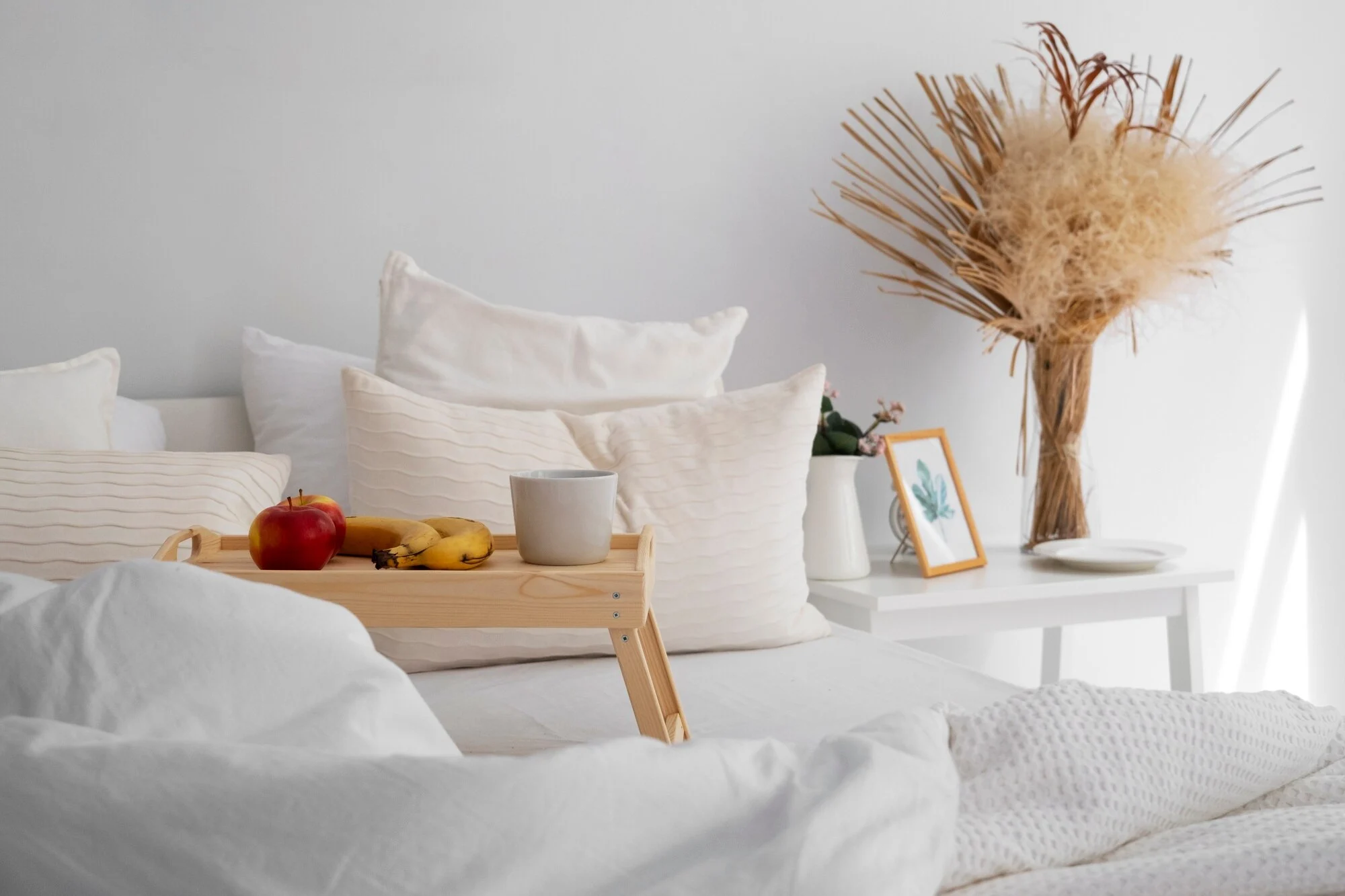 A cozy bedroom scene featuring a bed covered in white linens, with a wooden tray holding an apple, bananas, and a cup, surrounded by decorative pillows and a decorative dried flower arrangement on a side table with a picture frame.