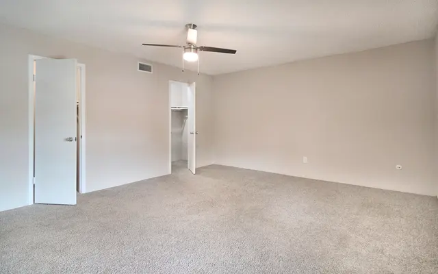 A spacious, empty room with gray carpeting, a ceiling fan, and light beige walls. There is a closet entrance visible on the right, and an open door on the left leading to another room.