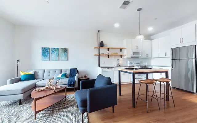Modern Apartment Interior Design A modern apartment interior featuring a light-filled living area with a gray sectional sofa, black chairs, and a wooden table. The kitchen is equipped with white cabinetry and stainless steel appliances.