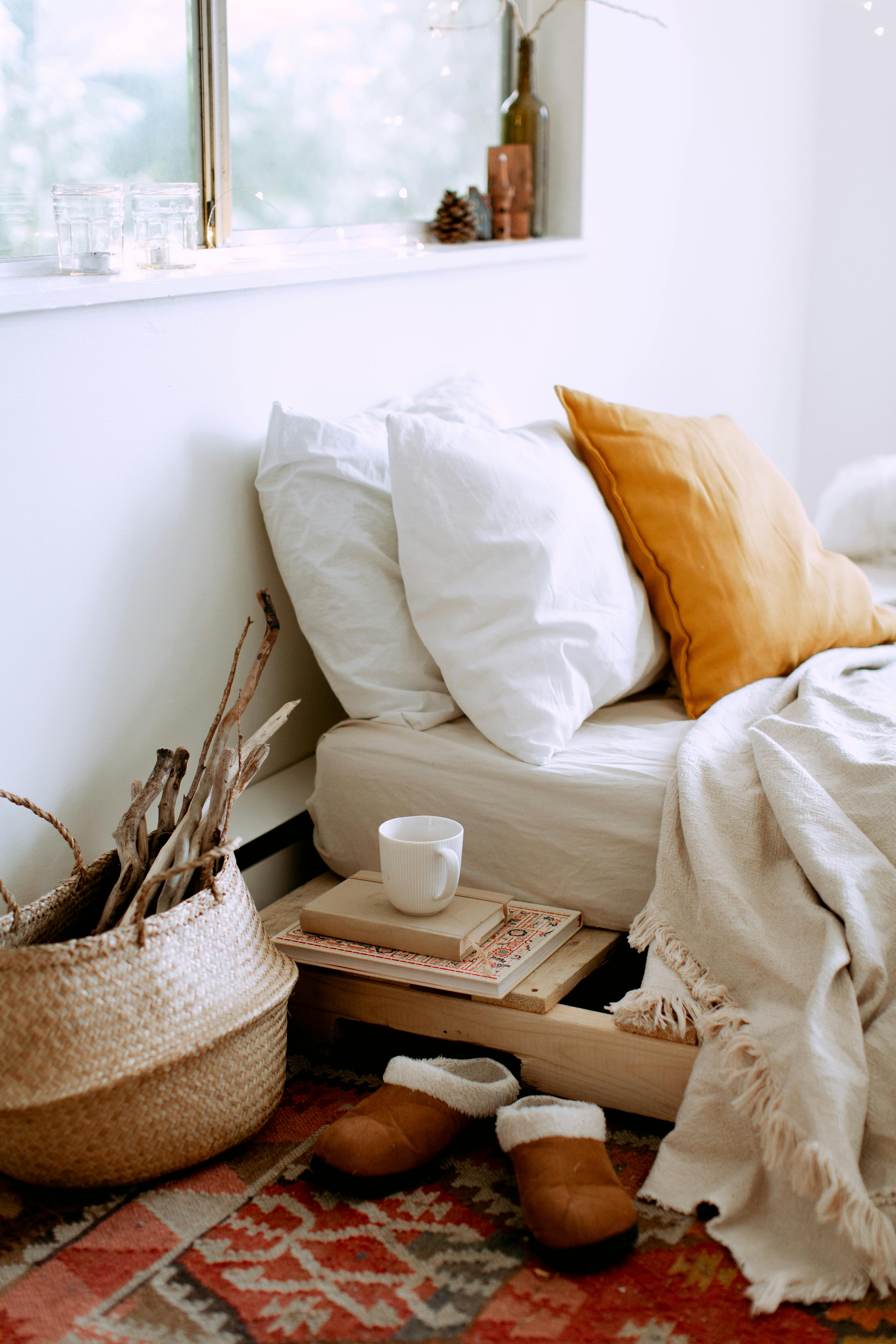 Cozy Bedroom Nook A cozy bedroom nook featuring a made bed with white pillows and a mustard yellow throw pillow. A woven basket holds sticks, and a stack of books sits on a wooden nightstand next to a white mug. A patterned rug is visible on the floor.
