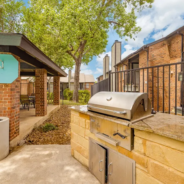 An outdoor barbecue area with a grill and seating beneath a covered structure, surrounded by green trees and a blue sky with clouds.