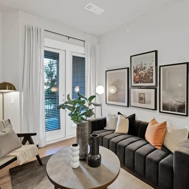 Chic Modern Living Room A modern living room showcasing a stylish sofa, decorative pillows, a coffee table, and wall art. The room features natural light from large windows with sheer curtains and a plant on the table.