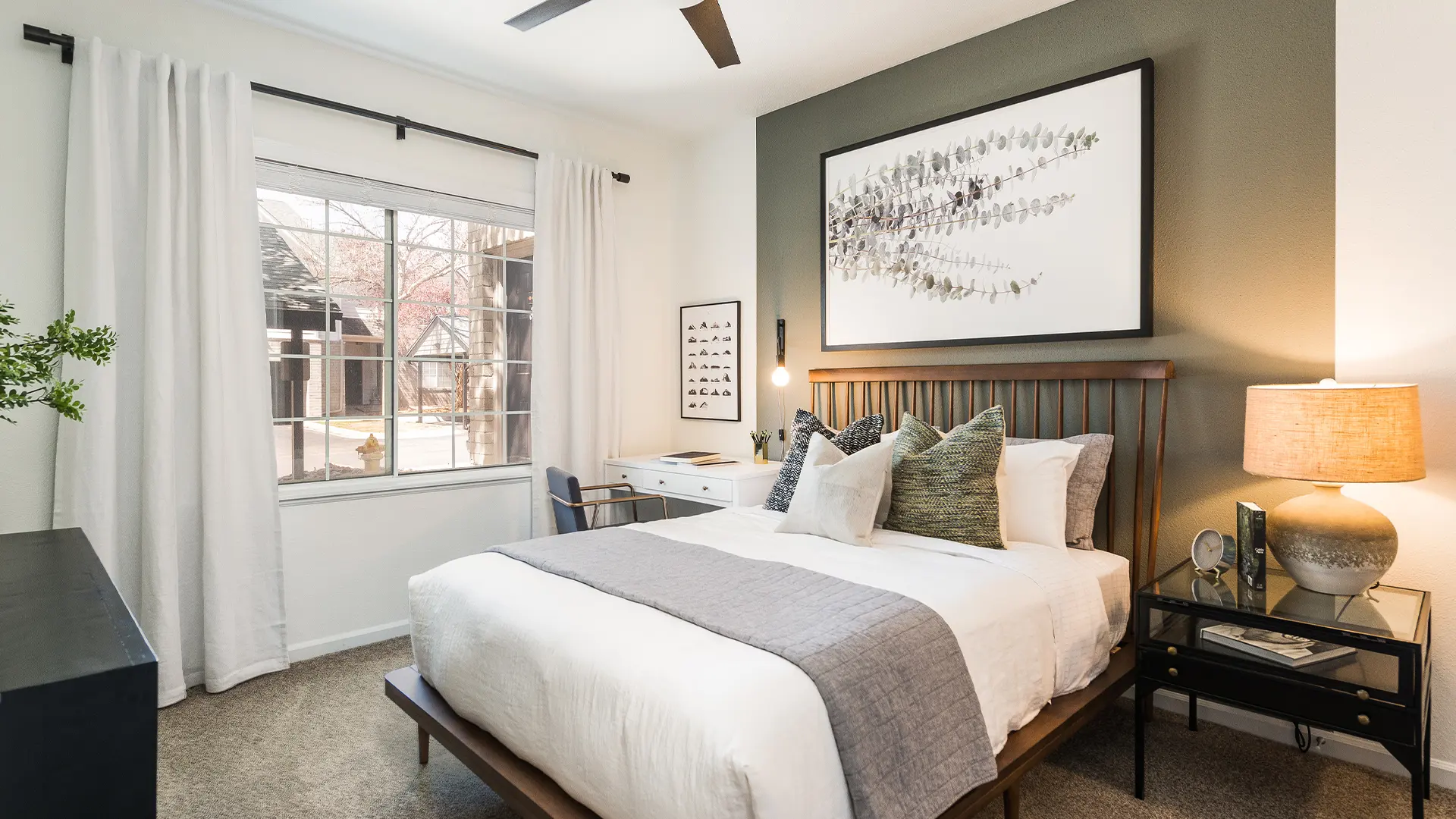 A modern bedroom featuring a queen-size bed with a white comforter, decorative pillows, and a gray throw blanket. The room has a dark green accent wall and a large window with sheer curtains, allowing natural light to enter. There is a lamp on a nightstand and a desk beside the bed, with artwork hanging above the bed.