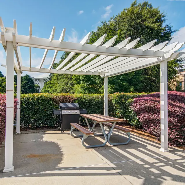 An outdoor space featuring a white pergola above a picnic table with a grill nearby, surrounded by colorful bushes and trees.