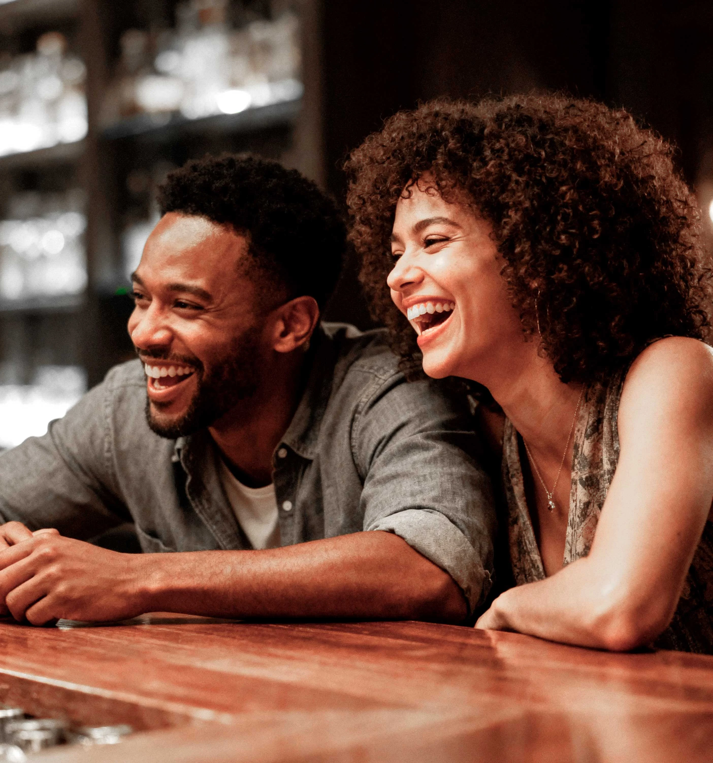 Two friends laughing together at a bar, with a wooden counter and blurred background bar shelves filled with drinks.