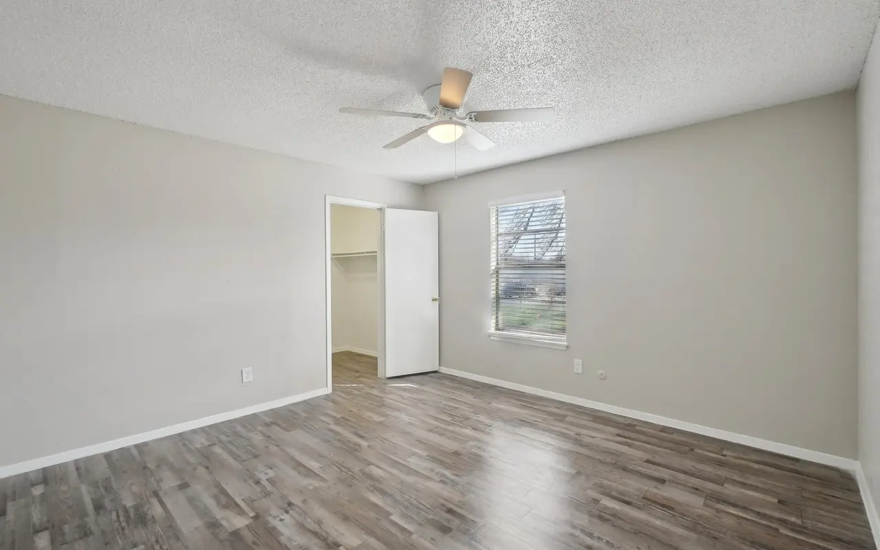 A spacious empty bedroom with a ceiling fan, light-colored walls, and wooden flooring. There is a closet with an open door and a window allowing natural light to enter.