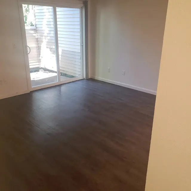 An empty living room with a wooden floor and large sliding glass doors leading to a small outdoor area. The walls are painted a light color and there is natural light coming in from the doors.
