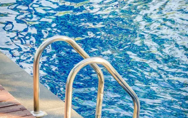 A silver pool ladder leading into a blue swimming pool, with reflections on the water surface and wooden decking visible.