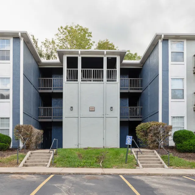 Front view of a three-story apartment building with balconies on each side, surrounded by grass and small bushes.