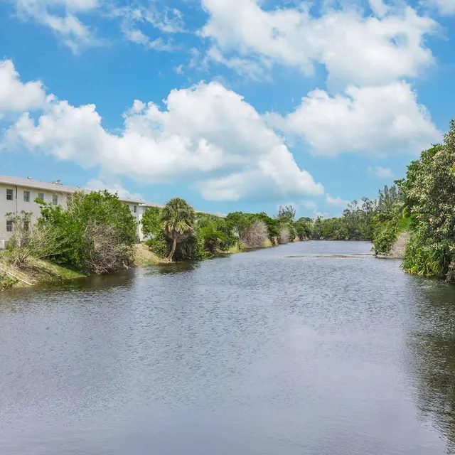 A tranquil river view surrounded by lush greenery, with a blue sky and fluffy clouds above. There are residential buildings visible in the background.