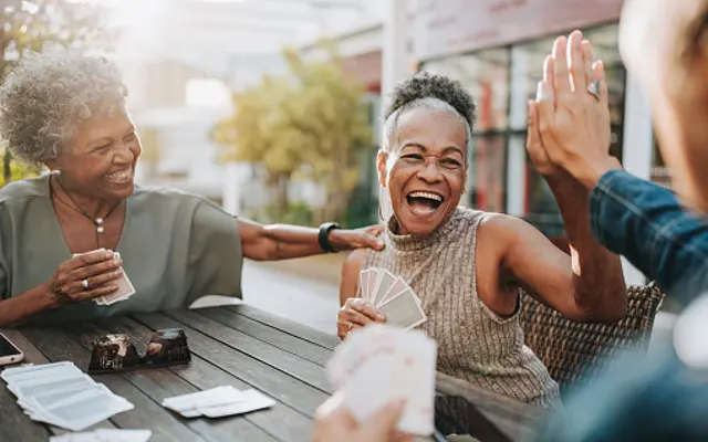 Joyful Card Game Among Friends Two women laughing and enjoying a card game outdoors, with a third person partially visible. One woman is holding cards and the other is playfully touching her shoulder.