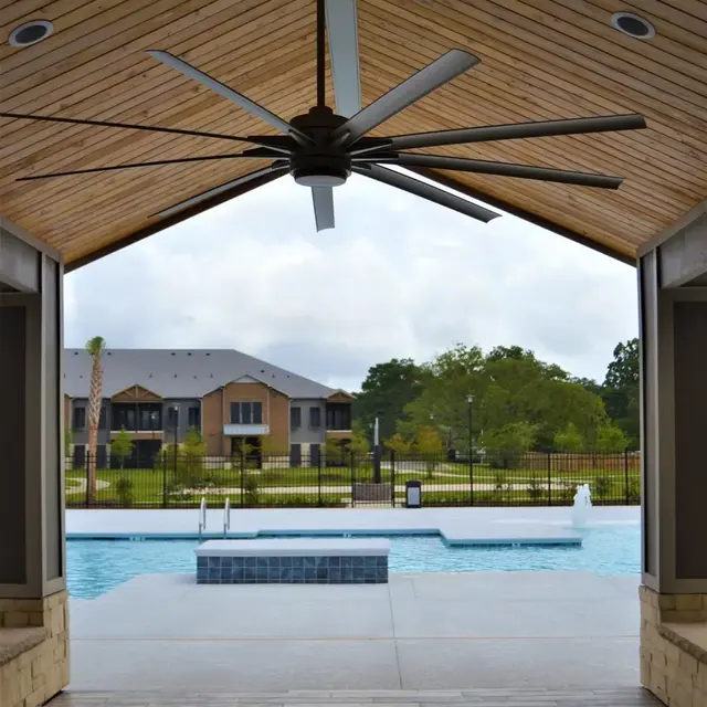 An interior view of a spacious structure with a wooden ceiling, looking out towards a swimming pool surrounded by greenery and residential buildings in the background.