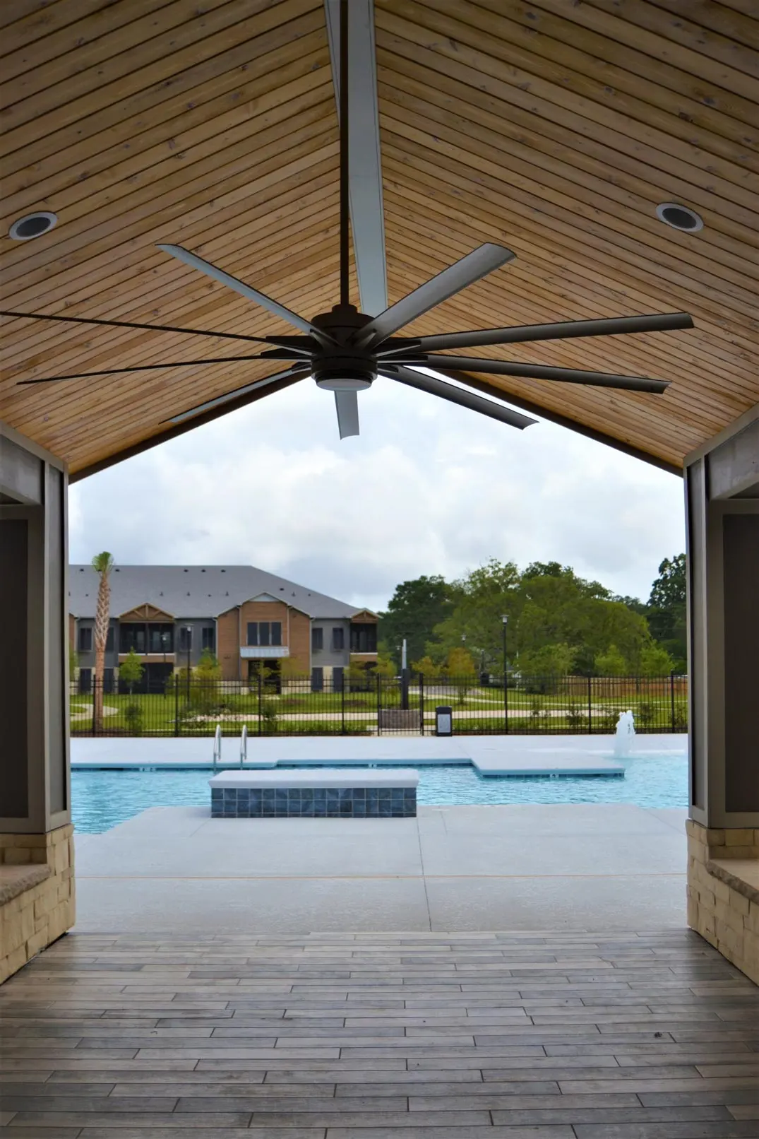 An interior view of a spacious structure with a wooden ceiling, looking out towards a swimming pool surrounded by greenery and residential buildings in the background.