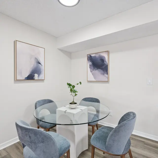 A small dining area featuring a round glass table surrounded by four soft blue chairs. On the wall, there are two art pieces with abstract designs in blue and white. A small potted plant sits in the center of the table.