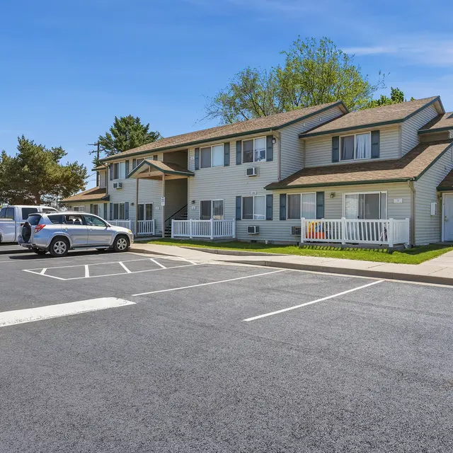 Apartment Building and Parking Area A two-story apartment building with a beige and white exterior, surrounded by grassy areas and trees, featuring a parking lot with several vehicles.