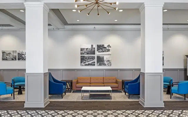 A modern lobby featuring blue chairs, a wooden sofa, and a coffee table under a contemporary chandelier.