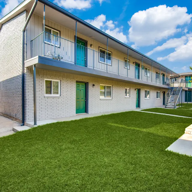 Exterior view of an apartment complex featuring two floors with green doors, surrounded by a manicured lawn and trees.