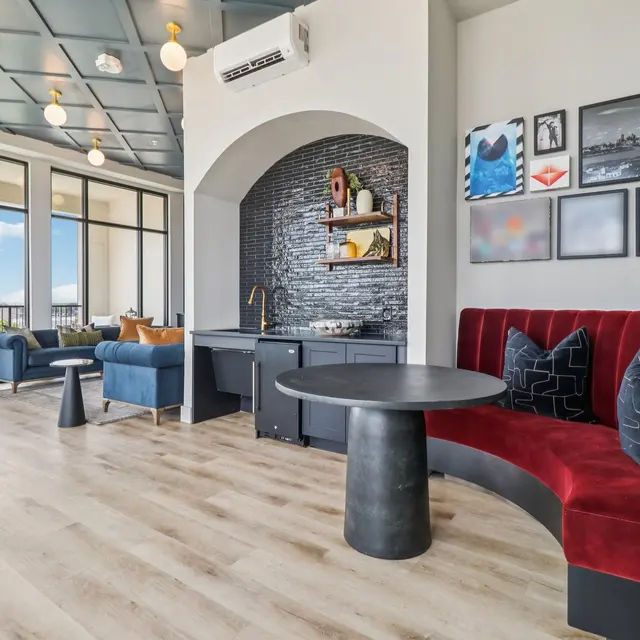 A modern lounge area featuring a red curved banquette, a round black table, and blue armchairs. The room has large windows allowing natural light and displaying outdoor views. Decorative frames hang on the walls, and there is a shelf with decorative items on one side.