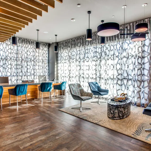 A stylish modern office lounge with wooden flooring and large patterned windows. It features a reception desk, blue chairs, and various seating arrangements including comfortable armchairs and a coffee table.