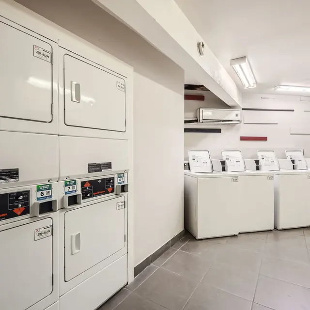 A clean and modern laundry room featuring stacked washer and dryer units on the left and several front-loading washing machines on the right, with bright overhead lighting and tiled flooring.