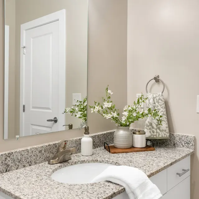 A modern bathroom with a granite countertop, a mirror, and decorative items including flowers in a vase and soap dispensers.