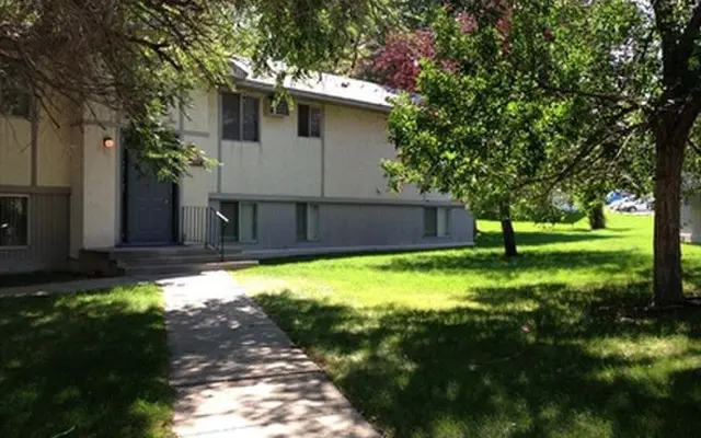 Exterior view of Twin Cedars Apartments in Helena, Montana, with lush green lawn, mature shade trees, and a welcoming walkway to the entrance