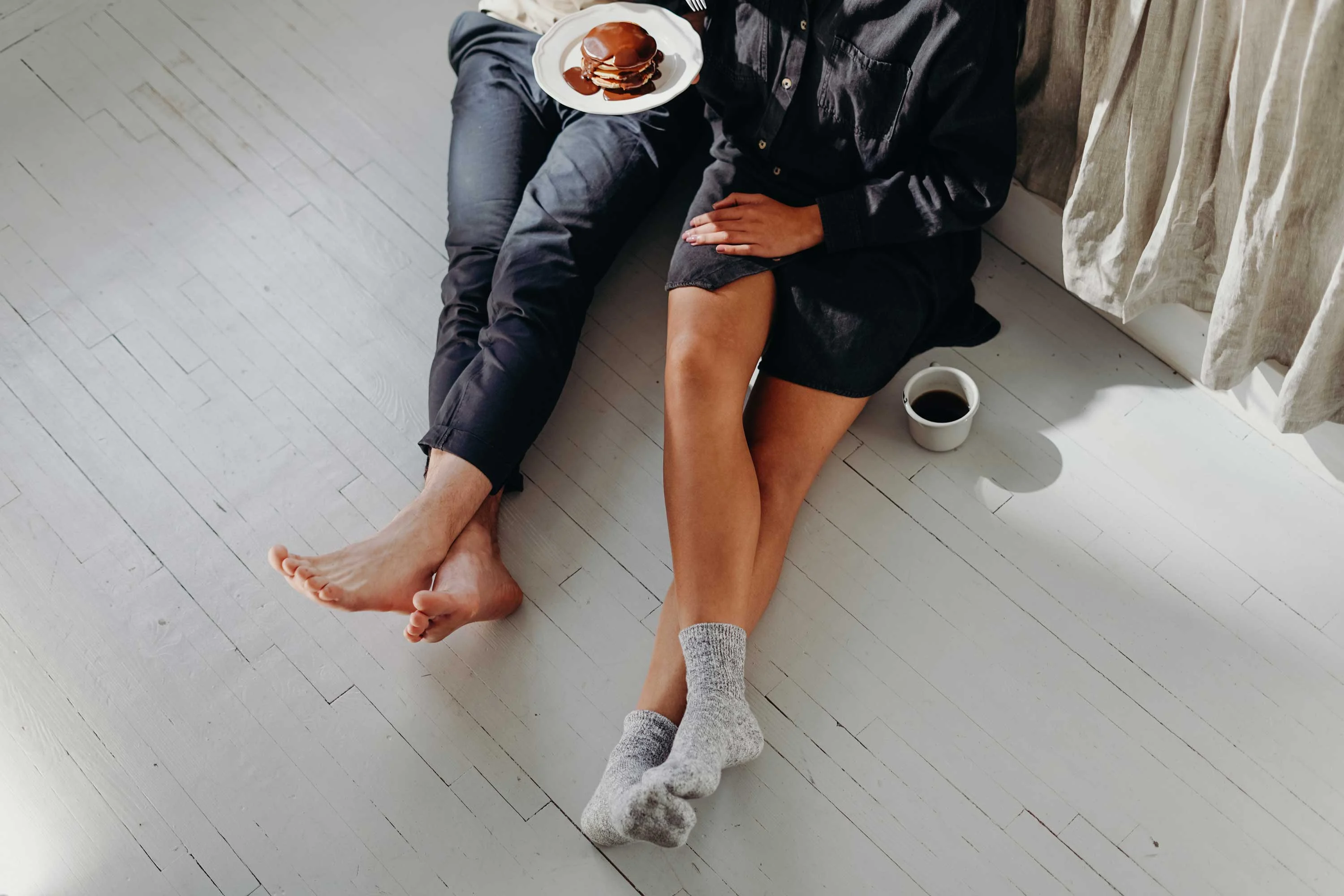 A couple sitting on a wooden floor, one holding a plate of pancakes and the other wearing cozy socks, with a cup of coffee nearby.