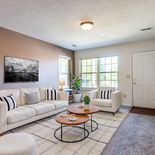 A modern living room featuring a white sofa with black and white striped cushions, a round coffee table, and a plant in the corner. There is a large window allowing natural light, and a door leading outside. The walls are painted in a light brown color, and there is a piece of artwork hanging on the wall.