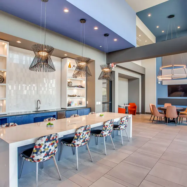 A modern community space featuring a long table with colorful chairs, shelves displaying decorative items, and distinct pendant lighting.