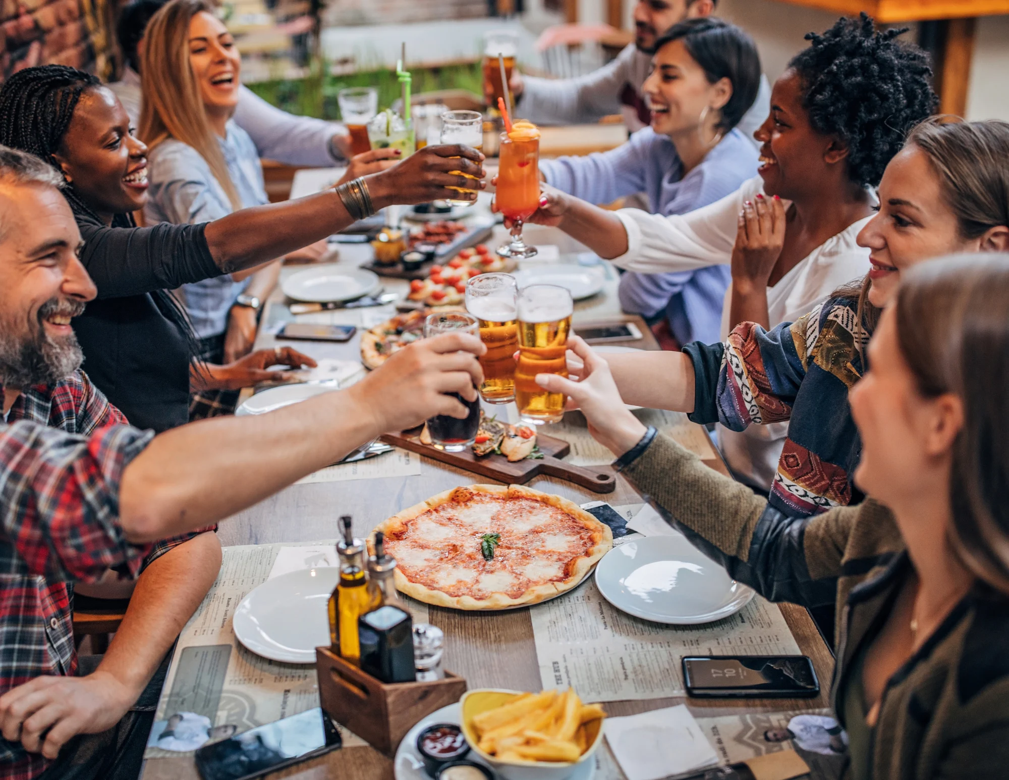 A group of diverse friends enjoying a festive meal at a long table filled with food and drinks, celebrating together with raised glasses.