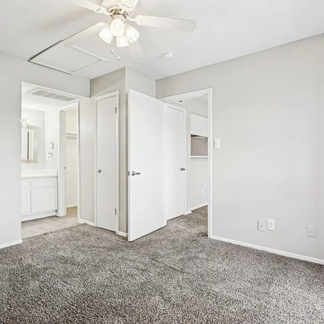 A spacious, empty bedroom with carpeted flooring, featuring a ceiling fan and two closed doors leading to adjacent rooms. The walls are painted in a light shade and there's a mirror visible in a nearby bathroom.