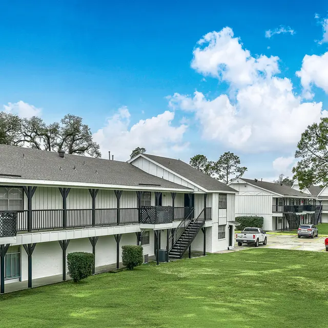 View of a multi-unit apartment complex with a green lawn surrounded by trees and a clear blue sky.
