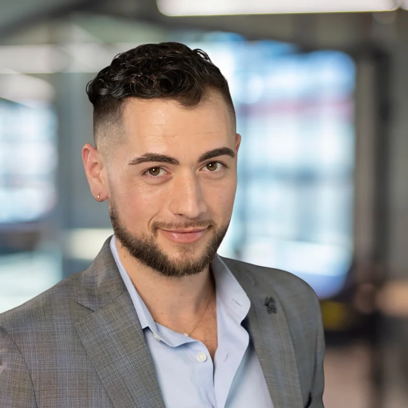 A young man with short, curly hair and a well-groomed beard, wearing a light blue shirt and a grey blazer, smiling confidently in a modern office setting with soft lighting and blurred background.