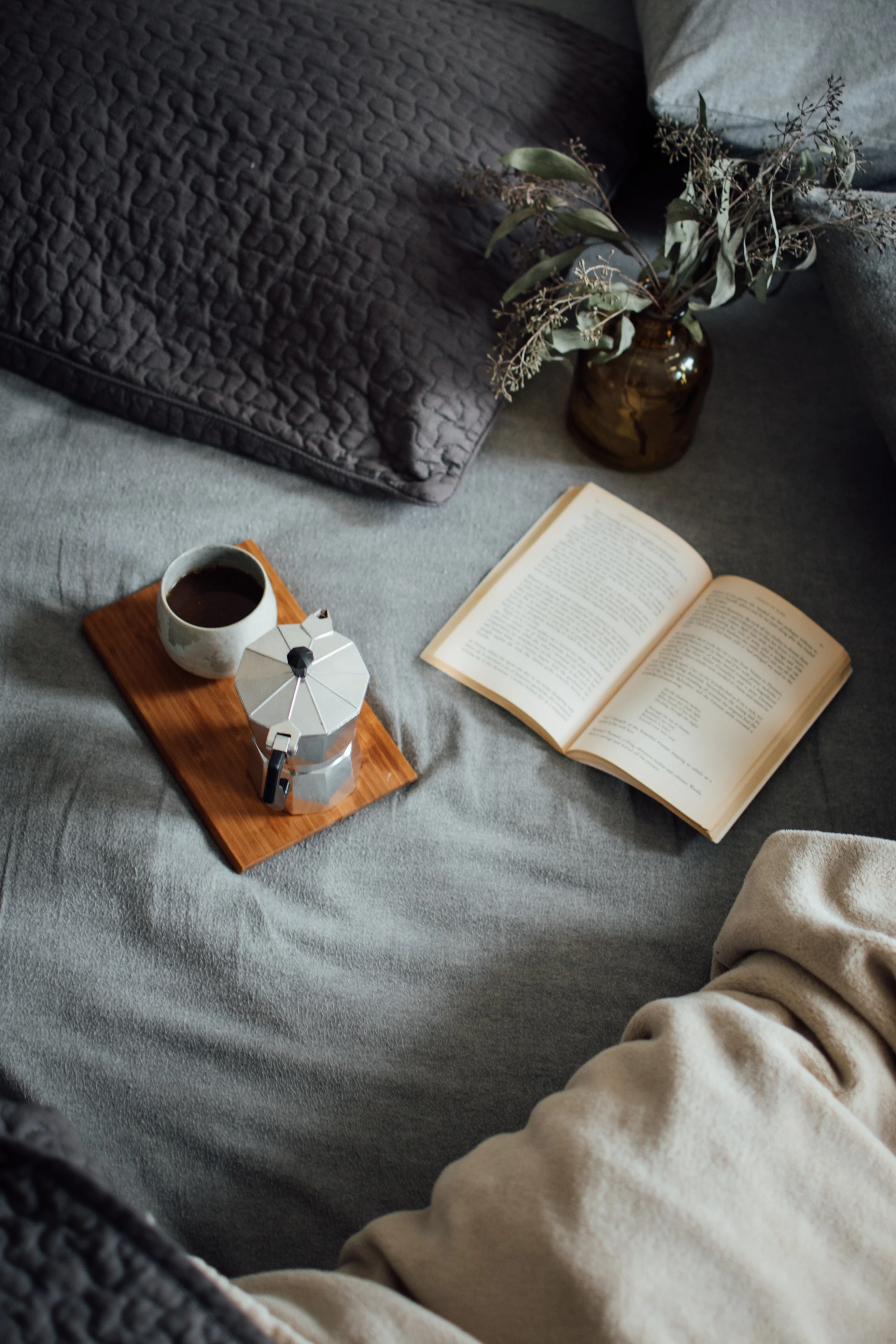 A cozy setup featuring a coffee maker, a cup of coffee, an open book, and dried flowers on a wooden tray, placed on a bed with gray linens and pillows.