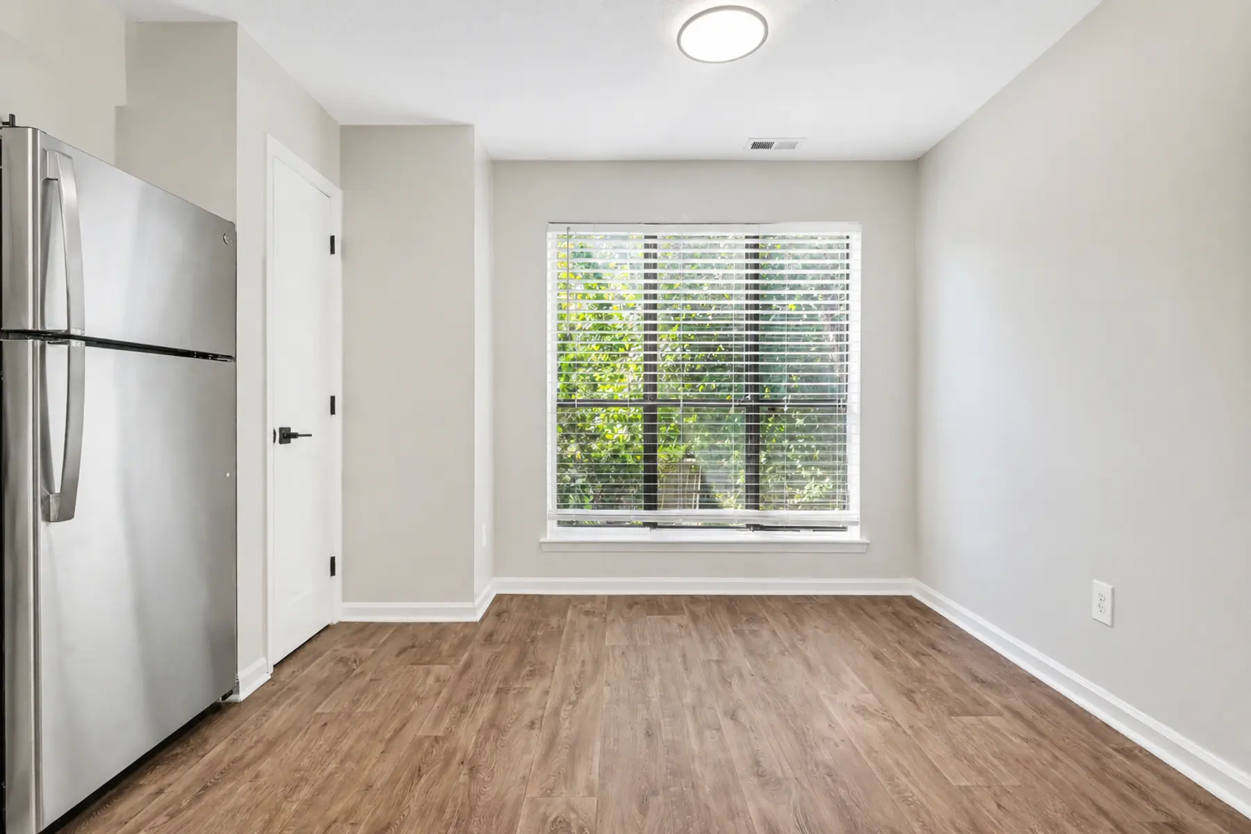 An empty room featuring a stainless steel refrigerator, hardwood flooring, and a large window with blinds.