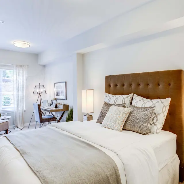 A cozy modern bedroom featuring a large bed with decorative pillows, a brown upholstered headboard, and bedside lamps. The room has a soft color palette and a small work area with a chair and desk visible in the background by a window.