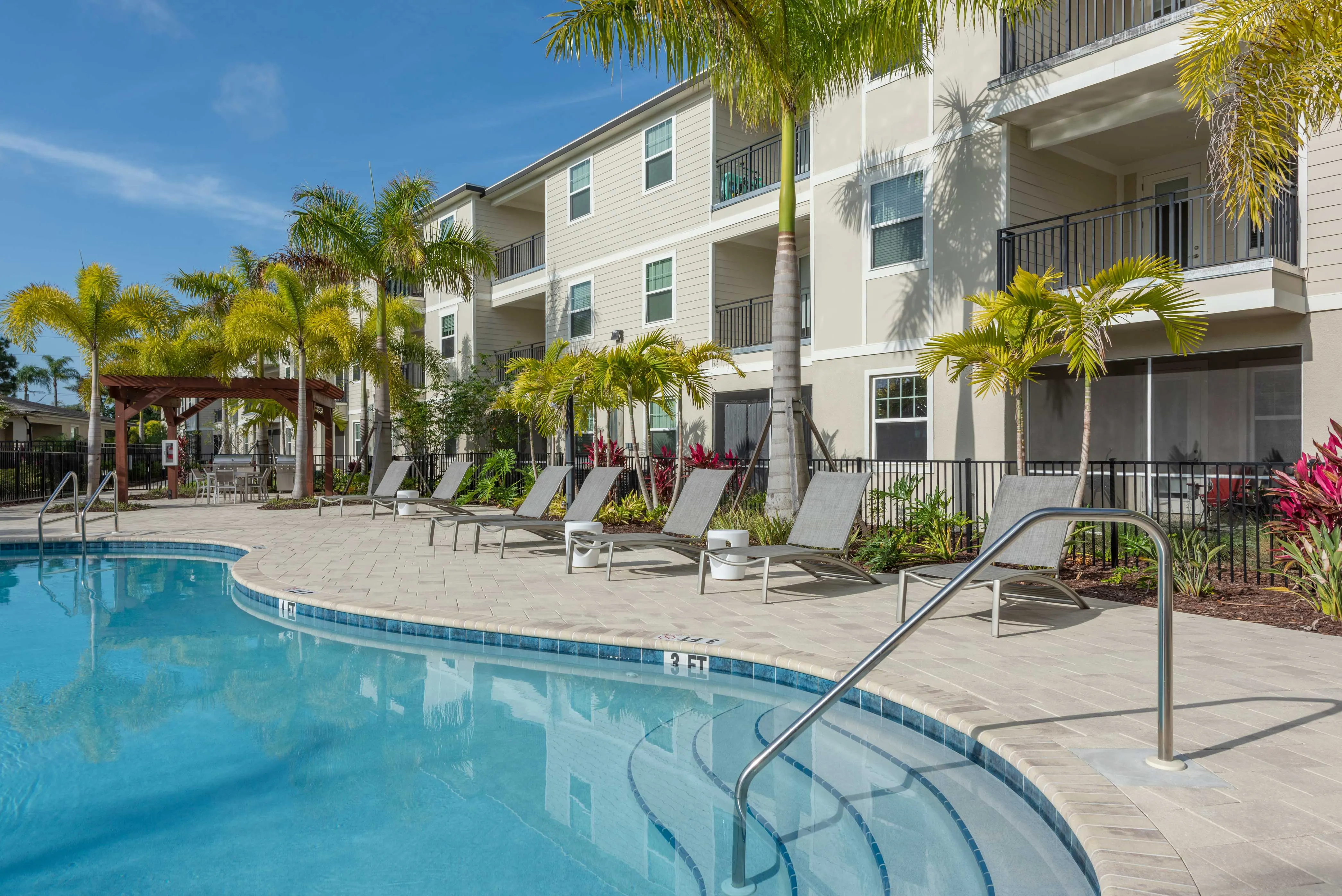 A spacious swimming pool area featuring lounge chairs surrounded by tropical plants and palm trees, with a backdrop of multi-story apartment buildings under a clear blue sky.
