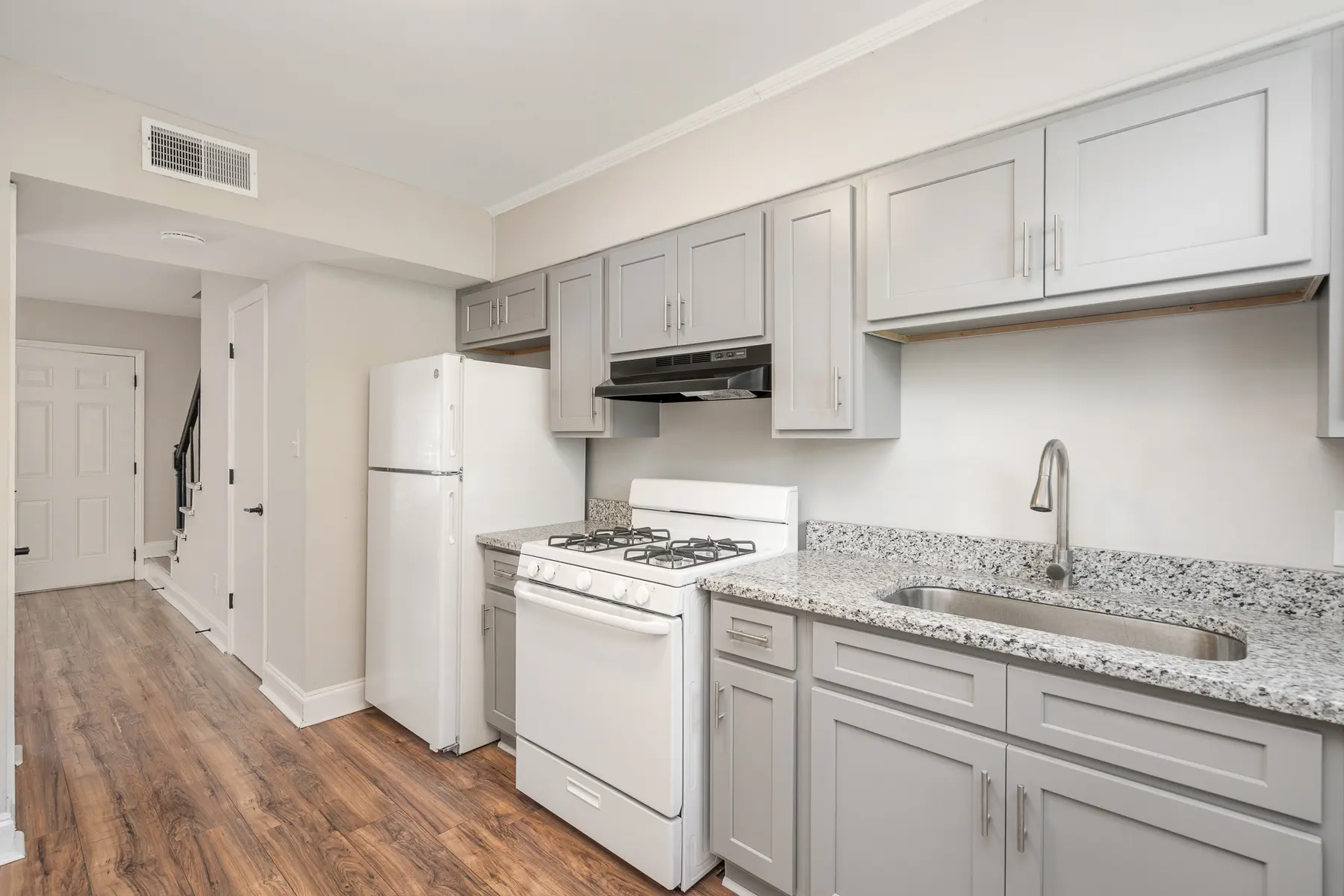 A modern kitchen featuring gray cabinetry, granite countertops, and a white gas stove. There's a refrigerator present, and the flooring is a warm wood tone. A doorway is visible in the background, leading into another area of the home.