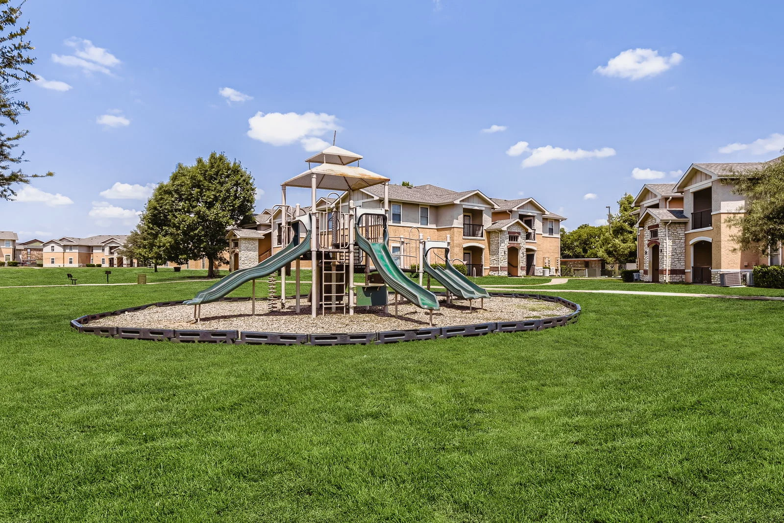 A playground with two green slides located on a grassy area surrounded by residential buildings.
