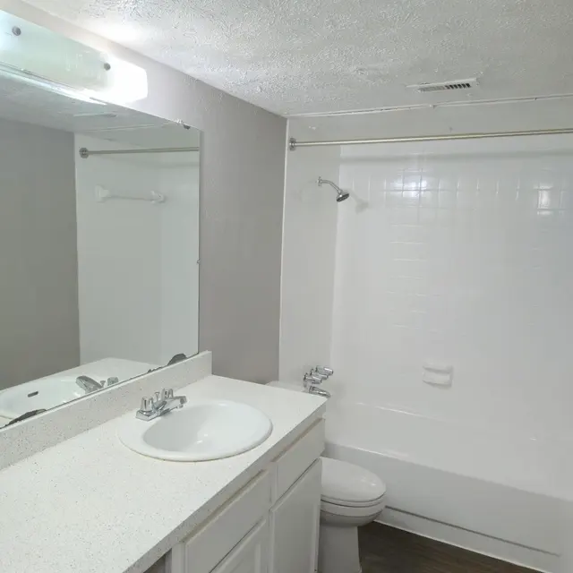 A clean, modern bathroom featuring a white countertop with a sink, a large mirror, and a bathtub with shower. The walls are painted in a light color and the flooring is dark wood.