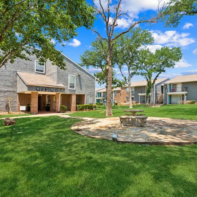 A landscaped view of an apartment complex with a grassy area, mature trees, and a stone circle.