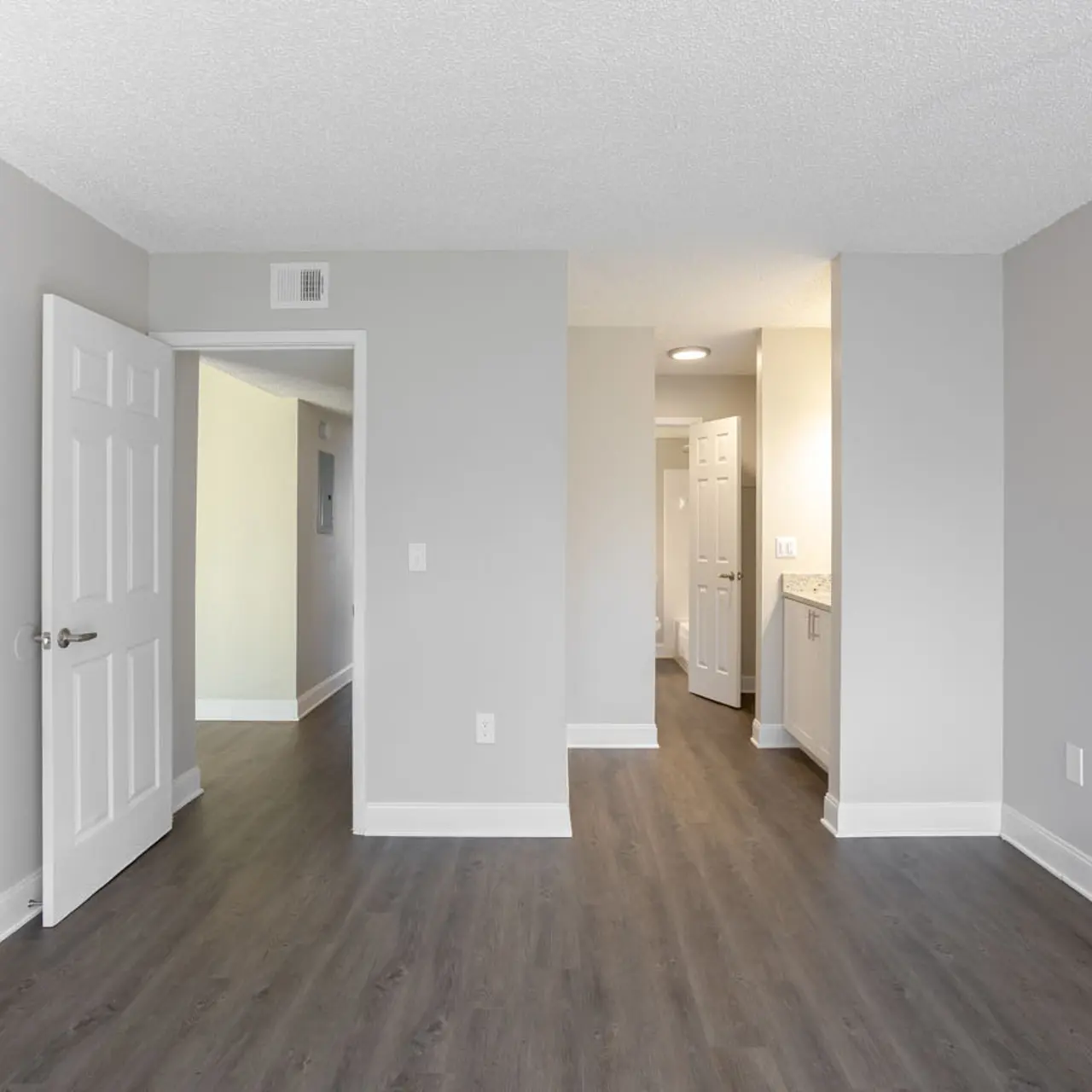 An empty room with gray walls and wooden flooring, featuring a white door on the left and an open doorway leading to another area. A bright hallway with light fixtures is visible in the background.