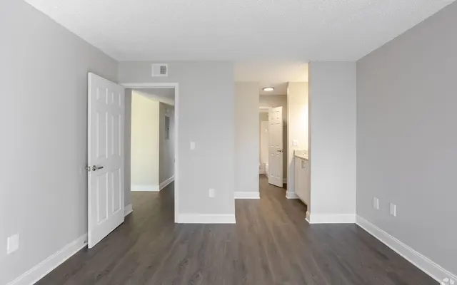 Modern Apartment Interior An empty room with gray walls and wooden flooring, featuring a white door on the left and an open doorway leading to another area. A bright hallway with light fixtures is visible in the background.