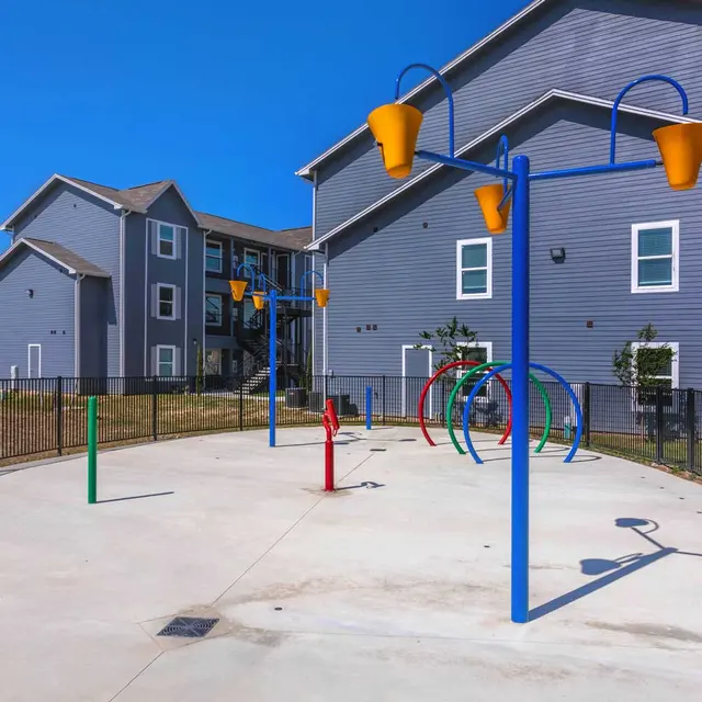A colorful playground area with a water spray feature surrounded by a black fence and gray apartment buildings in the background under a clear blue sky.