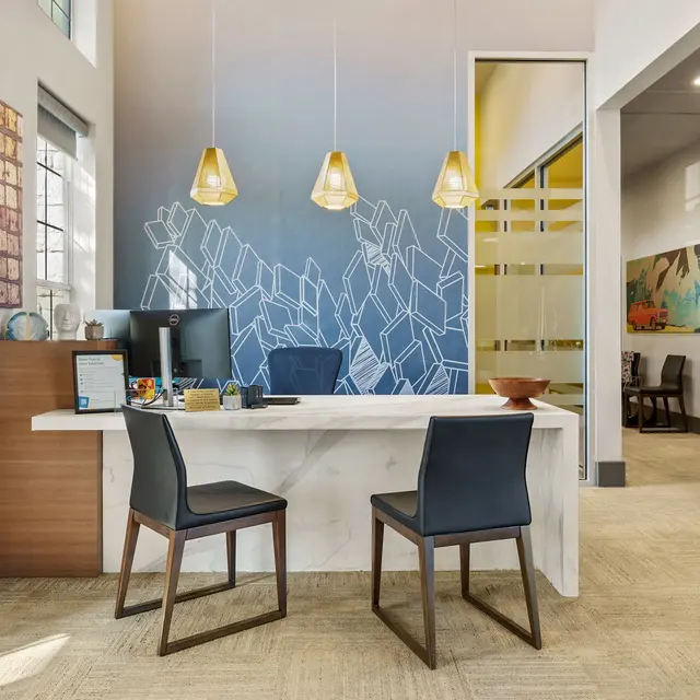 A modern office reception area with a stylish white desk, two black chairs, and pendant lights hanging above. The wall features a gradient blue design and geometric shapes, with artwork displayed nearby.